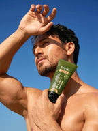Shirtless Tiger Shroff outdoors under a blue sky, shielding his eyes from the sun with one hand while holding a tube of PROWL Daily Sunscreen against his chest.